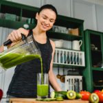 Happy young slim girl making fresh organic smoothie using blender in the kitchen