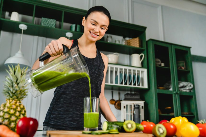 Happy young slim girl making fresh organic smoothie using blender in the kitchen